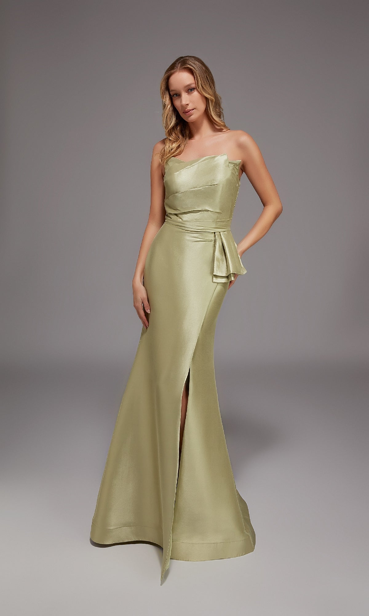 A woman poses in a studio wearing the Alyce 27806 long formal dress in honeydew, featuring a one-shoulder design, side slit, and waist sash.